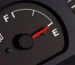Conductor preocupado mirando el tablero de su vehículo mientras carga combustible; representa el aumento en el consumo de bencina y los problemas mecánicos que pueden causarlo.